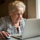 Elderly woman sitting at the table and types on laptop.