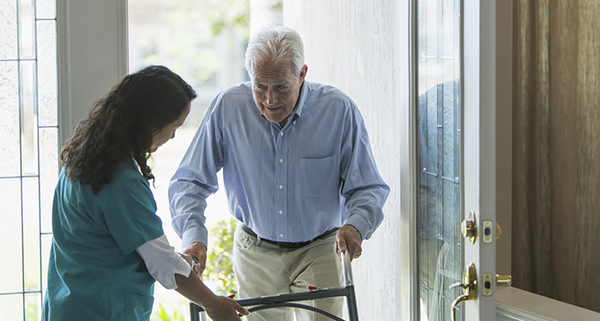 A home healthcare worker, a mature woman of Pacific Islander ethnicity, helping a senior man with a walker enter his house through the front door. She is holding the walker, directing him to watch is step.